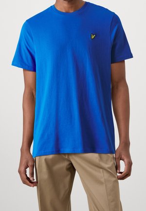 Uomo che indossa una maglietta a maniche corte di un vivace blu uniforme con un piccolo logo giallo di un uccello sul petto e pantaloni beige.