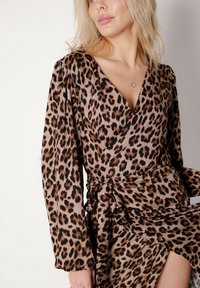 Sukienka kopertowa w panterkę z lekkiego materiału; posiada długie rękawy, głęboki dekolt w kształcie litery V oraz wiązany pas podkreślający boczne rozcięcie.