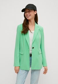 Mintgrüner Blazer mit tailliertem Design, einreihiger Knopfverschluss, zwei Fronttaschen und langen Ärmeln, kombiniert mit einer schwarzen Schirmmütze und einem weißen Oberteil.