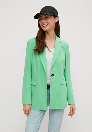 Muntgroene blazer met een op maat gemaakt ontwerp, enkele knoopsluiting, twee voorzakken en lange mouwen, gecombineerd met een zwarte pet en een witte top.