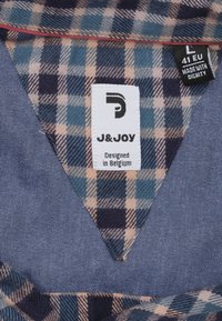 Ruitenkraag van een shirt met een blauw en beige ruitpatroon; heeft een effen blauwe stof eronder. Hangtag vermeldt "J&JOY," maat L, ontworpen in België.