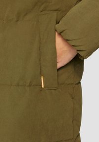 Olijfgroene pufferjack met een zichtbare nadenontwerp, voorzien van een zijzak en een klein merklabel op de stof. Zachte textuur.