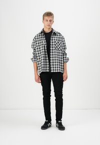 Young man standing wearing a black and white checkered shirt over a black t-shirt, black pants, and black sneakers with white stripes.