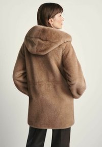 Kobieta ubrana w kapturem, jasnobrązowy futrzany płaszcz widziana z tyłu, zestawiona z czarnymi spodniami na jednolitym tle.