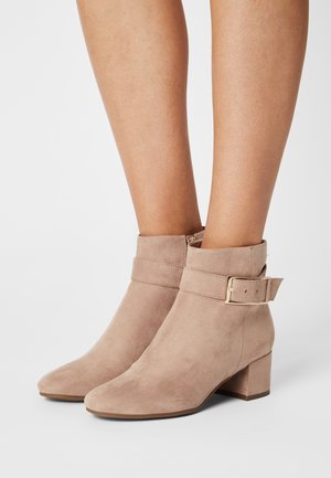 Bottes cheville en daim beige avec un talon bloc, dotées d'une large bride avec une boucle métallique et d'un bout arrondi. Texture lisse dans l'ensemble.