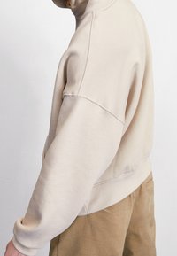 Personne portant un sweat-shirt beige oversize et un pantalon marron, vue de profil sur un fond blanc uni.