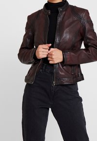 Person som bär en mörkbrun läderjacka över en svart polotröja och högmidjade svarta jeans, med händerna knäppta vid jackans dragkedja.