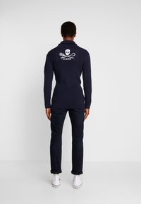 Marinblå ribbad tröja med hög krage och vitt dödskalle- och korsben-märke på baksidan. Kombinerad med mörka jeans och vita sneakers.