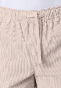 Pantalones de pana color beis con textura acanalada, cinturilla elástica y cordón ajustable. Presenta costuras mínimas y sin herrajes visibles.