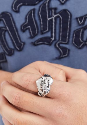 Bague en argent avec une forme de cœur portant le texte gravé "L'amour tue lentement". Le design comprend des accents décoratifs le long de l'anneau.