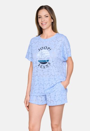 Femme souriante portant un haut à manches courtes bleu clair et un short avec un motif floral et le logo « JOOP! JEANS » sur le t-shirt.