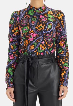 Femme portant un chemisier à manches longues et col montant à motifs floraux violets, verts, oranges et roses, rentré dans un pantalon en cuir noir taille haute.