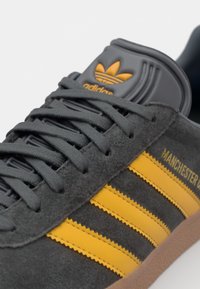 Gray suede sneaker with yellow stripes and logo. Features textured laces and a rubber sole with a brown accent. Classic design elements included.