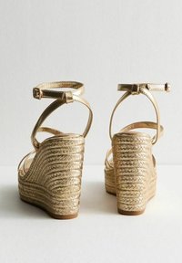 Sandalias de cuña doradas con textura de yute trenzado, correas en el tobillo y una parte superior brillante. La vista trasera resalta el diseño en capas de la cuña.