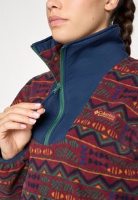 Pullover in pile con collo alto blu e corpo a motivo rosso vivace con forme geometriche. Cerniera verde e logo Columbia visibili.