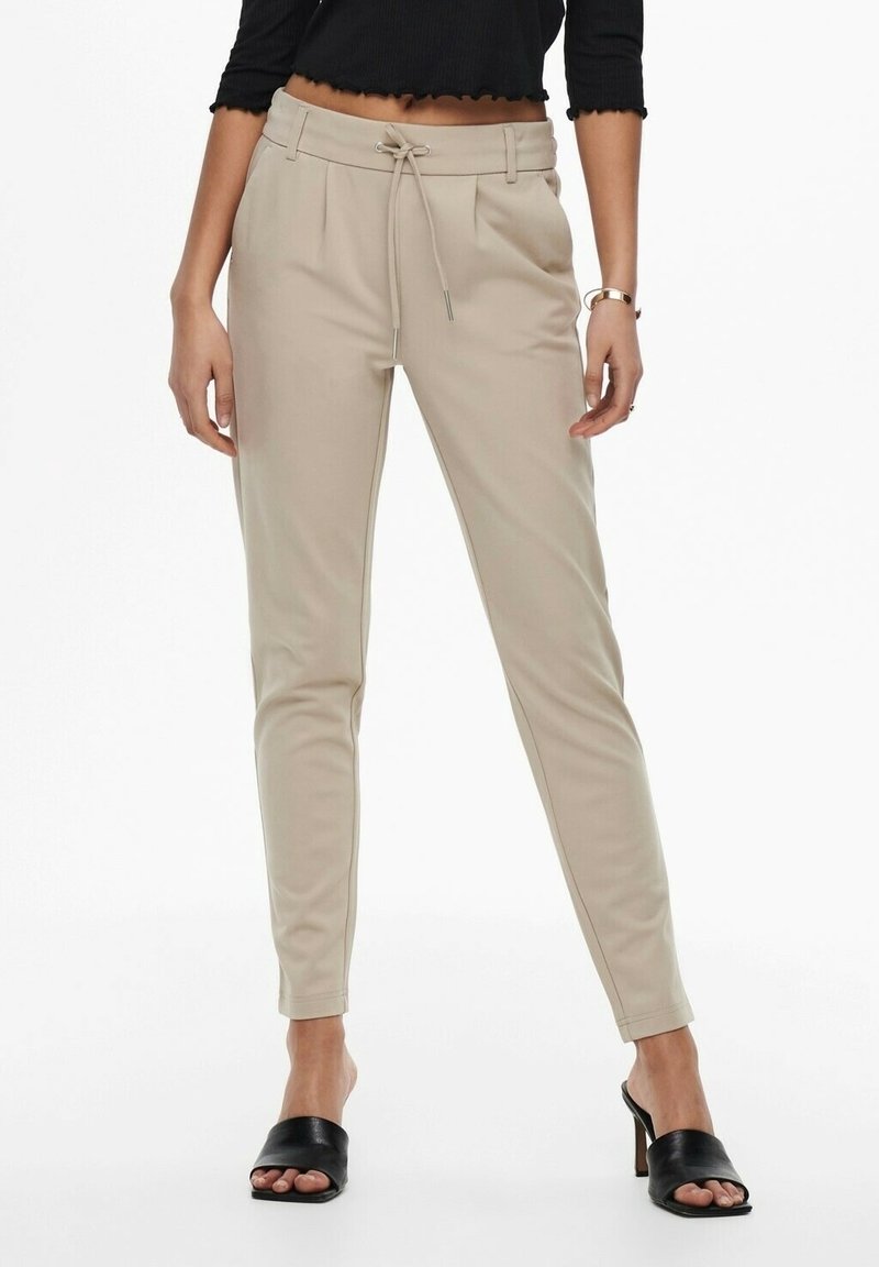 Femme portant un pantalon beige slim avec taille à cordon et des sandales à talons ouvertes noires, debout devant un fond blanc.