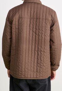 Mężczyzna w brązowej pikowanej kurtce, pokazany z tyłu, z pionowym przeszyciem i elastycznymi mankietami, w połączeniu z ciemnymi spodniami.