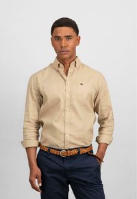 Tan overhemd met lange mouwen en knopen, voorzien van een borstzak en geborduurde details. Gekozen in combinatie met marineblauwe broek en een gevlochten leren riem.
