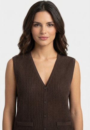 Mujer con cabello ondulado oscuro hasta los hombros, vestida con un chaleco tejido marrón sin mangas con botones, sobre un fondo claro y liso.