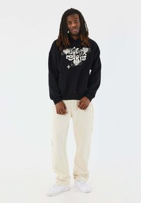 Sudadera negra con gráfico "Proud" en blanco, combinada con jeans color crema que tienen bordes deshilachados y zapatillas blancas.