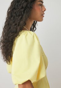 Gelbes Kleid mit Puffärmeln aus glattem Stoff mit Raffung. Gestylt mit einer Perlenkette vor einem hellen, neutralen Hintergrund.