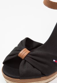 Sandale en tissu noir avec une bride en cuir marron clair et un détail de nœud sur une semelle style espadrille avec des coutures blanches.