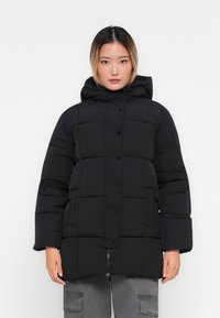 Mujer vestida con una chaqueta de invierno acolchada negra con capucha, botones de presión y cremallera, combinada con pantalones cargo grises, de pie frente a un fondo blanco.