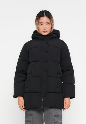 Mujer vestida con una chaqueta de invierno acolchada negra con capucha, botones de presión y cremallera, combinada con pantalones cargo grises, de pie frente a un fondo blanco.