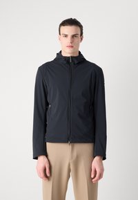 Black lightweight jacket with a zip front, hood, and side pockets. Smooth texture and tailored fit, paired with beige trousers.