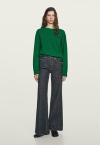 Maglione verde a maglia con maniche lunghe abbinato a jeans in denim scuro a vita alta e gamba larga. L'outfit presenta dettagli minimi.