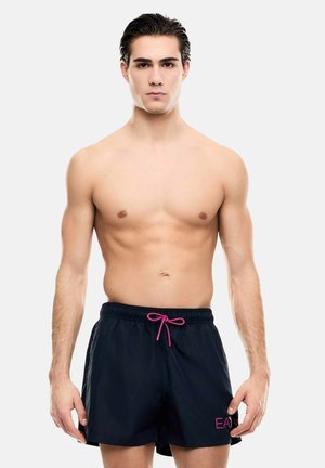 Giovane uomo con capelli bagnati che indossa pantaloncini da bagno neri con coulisse rosa e logo EA7, in piedi davanti a uno sfondo chiaro e uniforme.