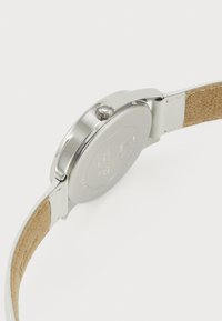 Sølvur med en glat metalcase, rund form, hvid læderrem og struktureret tan underside. Har en krone på siden.
