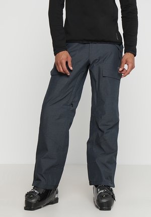Mann trägt schwarze Skischuhe, dunkelgraue Cargo-Schneehose und ein schwarzes Langarmshirt, steht vor weißem Hintergrund.