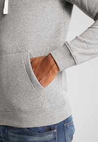 Grå hoodie med en ficka framtill, ribbade ärmslut och en texturerad yta. Tyget verkar mjukt och bekväm, lämpligt för avslappnad klädsel.