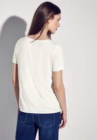 Bílý krátký rukáv tričko s kulatým výstřihem, hladkou texturou a jemným nařasením vzadu, kombinované s modrými džíny.