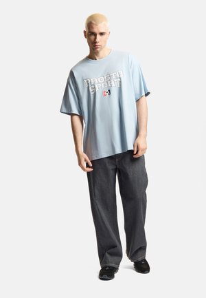 Młody mężczyzna o krótkich blond włosach, ubrany w jasnoniebieską koszulkę "Prosto Sport", luźne ciemne dżinsy i czarne trampki, stojący na białym tle.