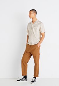Camicia beige a maniche corte con bottoni e due tasche sul petto, abbinata a pantaloni marroni arrotolati e sneaker a quadri bianco e nero.