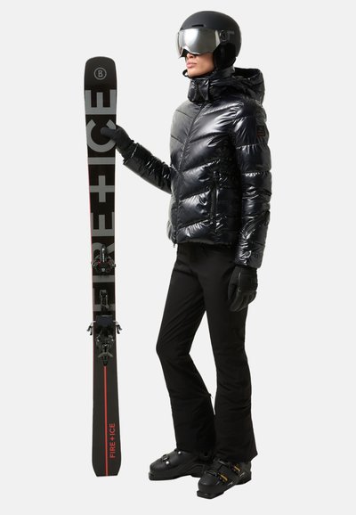 Skioutfit med en skinnende sort pufferjakke, sorte bukser, handsker og hjelm. Holder sorte ski mærket "FIRE + ICE."