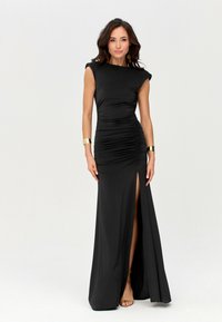 Woman wearing a black sleeveless ruched gown with a thigh-high slit, gold bracelets, drop earrings, and beige strappy heels, standing on white background.