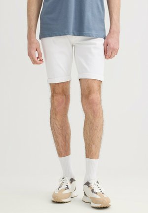 Hombre con pantalones cortos blancos remangados, zapatillas beige y blancas, calcetines blancos y una camisa azul, de pie contra un fondo liso.