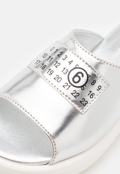 Sandale glissante métallique argentée avec un patch numéroté et un accent de cercle noir, dotée d'une surface texturée et brillante, et d'une semelle blanche.
