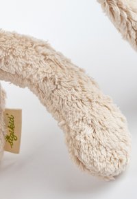 Beige plush toy with a soft, textured surface. Includes a fabric tag with green text. Features a curved, flexible shape resembling an animal limb.