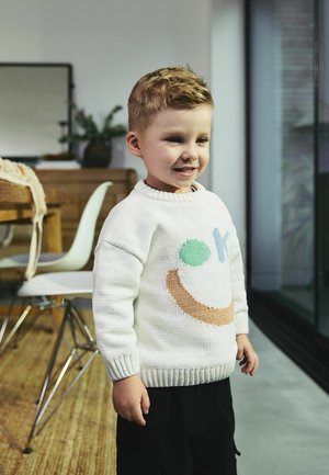 Weißer Strickpullover mit buntem Smiley-Gesicht Design, das blaue, grüne und braune Akzente aufweist. Rippkragen und -bündchen, kombiniert mit schwarzen Hosen.