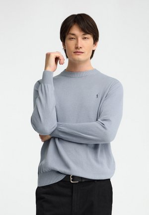 Hombre con cabello oscuro, vestido con un suéter azul claro y pantalones negros, posa con un brazo cruzado y la otra mano cerca de su rostro.