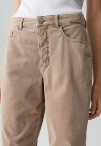 Pantaloni di cotone beige con una texture liscia, dotati di chiusura con bottone frontale, passanti per cintura e due tasche laterali. Visibile una semplice cucitura decorativa.