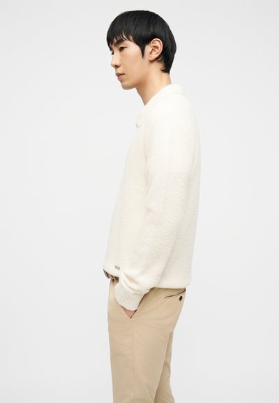 Νεαρός με μαύρα μαλλιά που φοράει λευκό πλεκτό πουλόβερ και μπεζ παντελόνι, στέκεται πλευρικά με τα χέρια στις τσέπες μπροστά σε απλό φόντο.