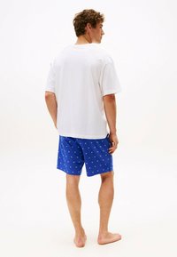 Maglietta bianca in cotone con maniche corte, abbinata a pantaloncini blu con un piccolo motivo geometrico bianco. A piedi nudi, vista da dietro.