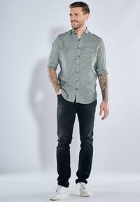 Hellgrünes Hemd mit Knopfleiste und hochgekrempelten Ärmeln, kombiniert mit schwarzen Jeans und weißen Sneakers. Verfügt über einen strukturierten Stoff und subtile Nähtedetails.