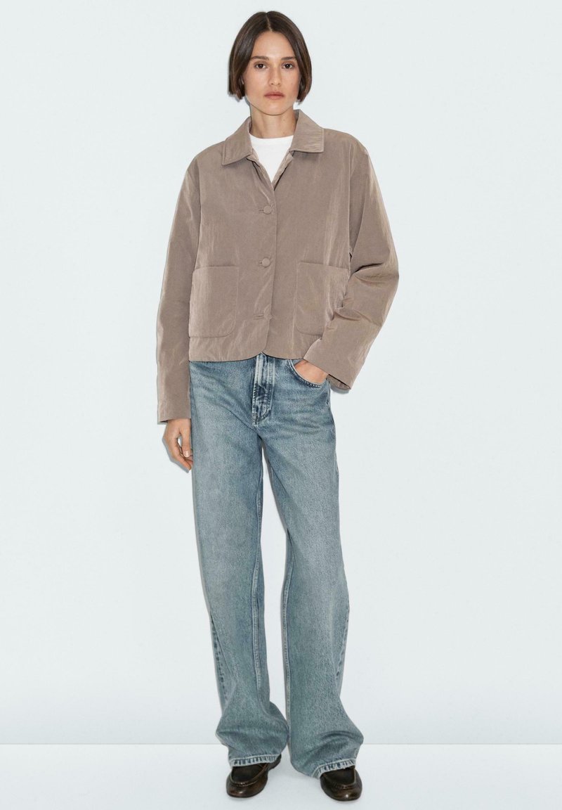 Geknipte, beige jas met een kraag, twee frontrijzakken en ronde knopen, gecombineerd met lichtblauwe wijde jeans en zwarte schoenen.