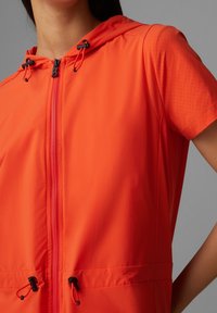 Giacca arancione con cappuccio e maniche corte, dotata di una zip frontale, vita con cordino e texture perforata sulle maniche.